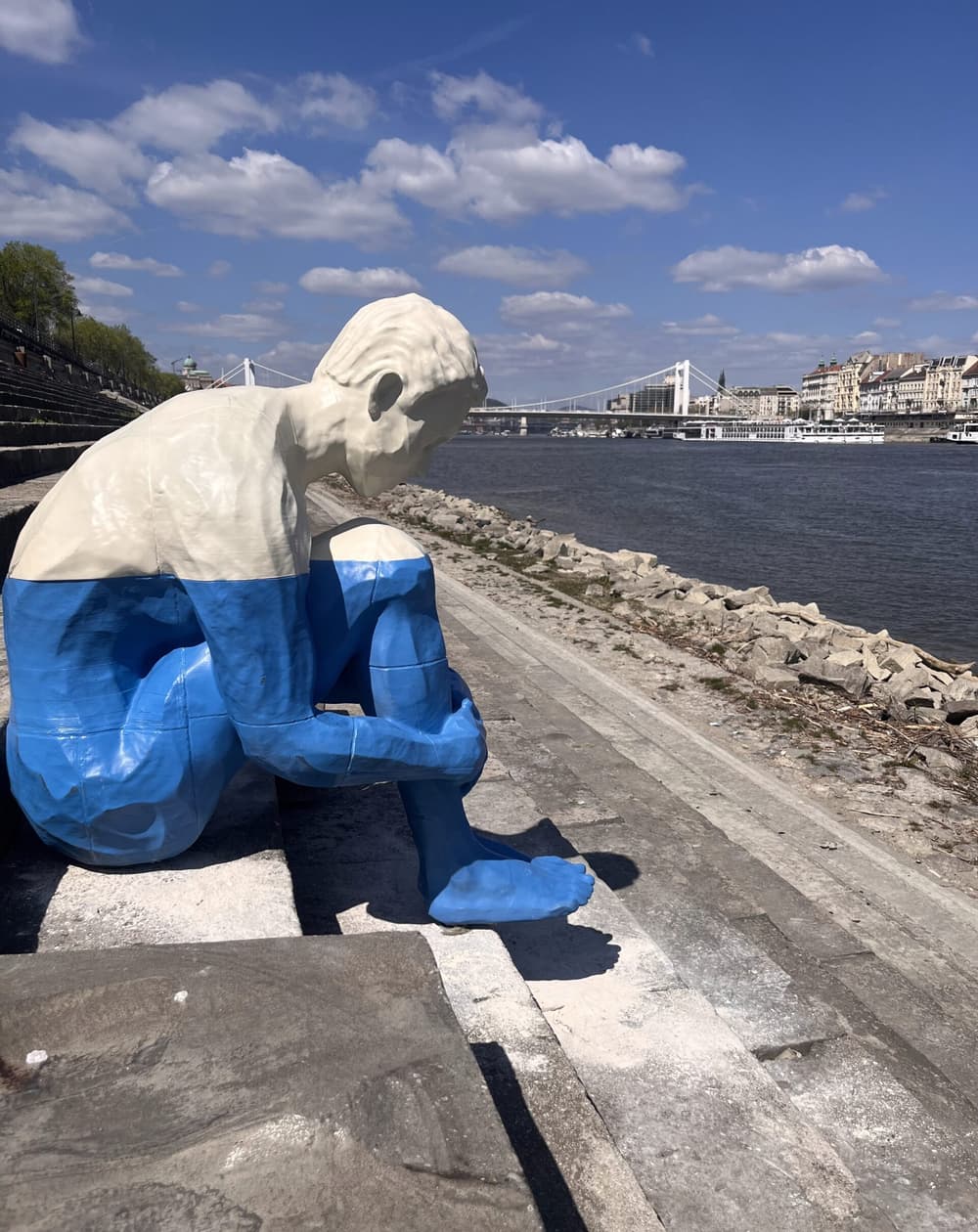 A cream-and-blue seated PETG sculpture on the Danube embankment steps, with a suspension bridge and the Budapest skyline in the background